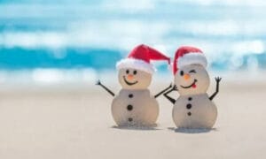 2 sand snowmen on the beach wearing santa hats.