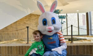 A smiling young child in a green shirt sits on a bench next to a large Easter Bunny mascot with white fur, pink ears, and blue overalls at Jackson College's Annual Easter Egg Hunt. The bunny has its arm around the child as they pose together. Kids are encouraged to say hi and take photos with the bunny mascot.