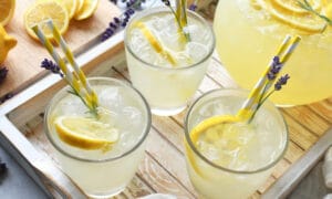 Lemonade cups sitting on a tray with garnish of lavender and lemon wedges on top with a straw all next to a pitcher of lemonade