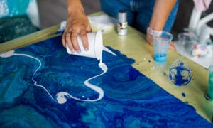A person pouring white paint onto a blue painting.